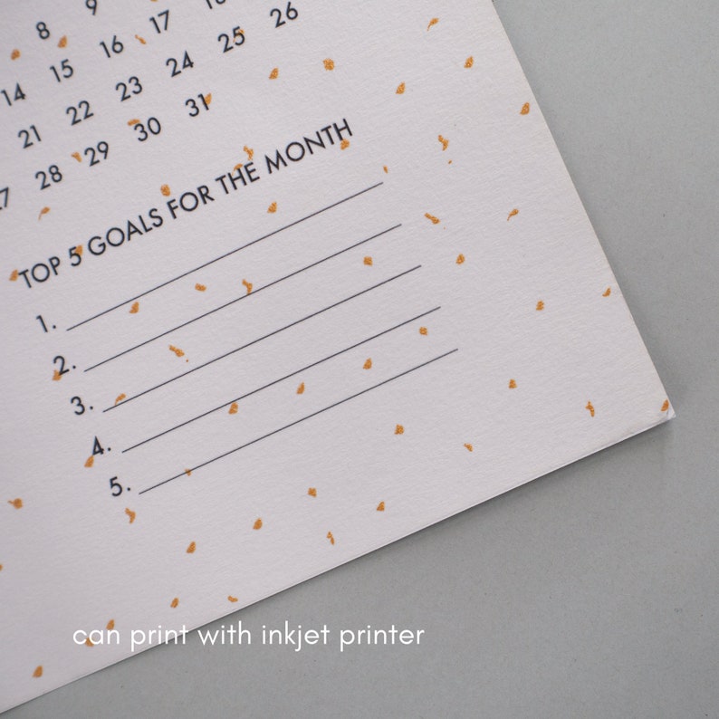 May include: A white calendar page with gold confetti printed on it. The calendar page has the numbers 8, 9, 14, 15, 16, 17, 21, 22, 23, 24, 25, 26, 7, 28, 29, 30, and 31 printed on it. The text "TOP 5 GOALS FOR THE MONTH" is printed at the top of the page. There are five blank lines below the text for writing goals. The text "can print with inkjet printer" is printed at the bottom of the page.
