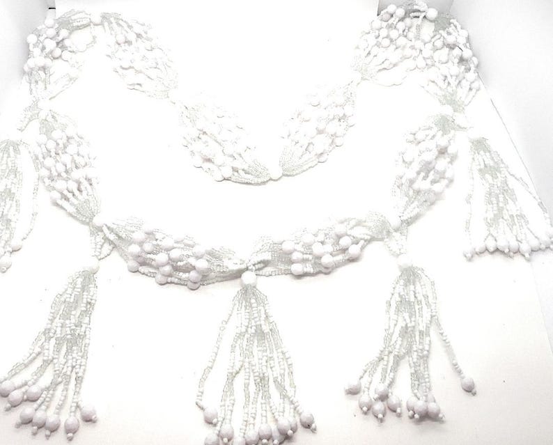May include: White beaded garland with multiple strands and tassels. The garland is made of small white beads and has a delicate, feminine look.
