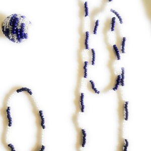 May include: A set of two necklaces made with white and purple beads. The longer necklace is a statement piece with a layered design. The shorter necklace is a simple bracelet.