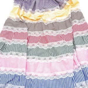 May include: A colorful gingham skirt with multiple layers of white lace trim. The skirt features alternating stripes of purple, yellow, brown, red, green, pink, and blue gingham.