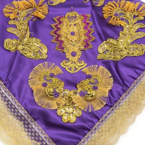 May include: A purple satin fabric square with gold and purple embroidery and sequins. The fabric has a gold lace trim around the edges.
