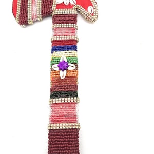 May include: A red and gold beaded axe with colorful stripes and shell accents. The axe is decorated with a purple gem and white shells.