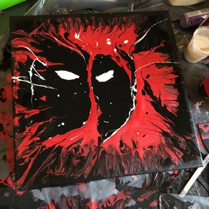 Pode incluir: Uma tela quadrada com um retrato abstrato de Deadpool. A obra de arte utiliza uma técnica de pintura fluida com uma paleta de cores ousada de vermelho, preto e branco. Os olhos são brancos, e o design geral é uma representação marcante do personagem.