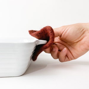 以下が含まれることがあります： 白いセラミック製のベーキングディッシュのハンドルの上に、茶色のウール製の鍋つかみを置いた手