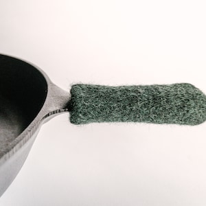 Puede incluir: Una sartén de hierro fundido negra con una cubierta de mango de lana verde oscuro.