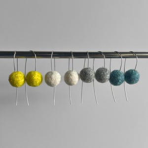 May include: A collection of colorful felt ball earrings in a variety of colors, including yellow, white, gray, and teal. The earrings are suspended from a silver bar, creating a visually appealing display. The earrings are held by a hand.