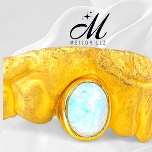 May include: A gold-coloured grill with a textured surface and an oval-shaped, light blue and white opal stone. The grill has the text "MEILGRILLZ" and a stylized "M" logo.