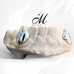 May include: A dental mold featuring two silver grillz. The grillz have a reflective surface with a cross design. The brand name "MEILGRILLZ" is displayed in black script above the mold.
