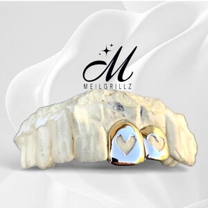 May include: A dental grill with a gold finish and heart-shaped cutouts. The grill is set on a white mold, with the brand name "MEILGRILLZ" in black script above. The background is a soft, white gradient.