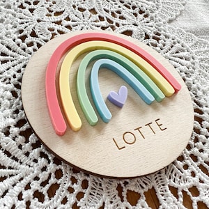 Könnte beinhalten: Ein Holz-Kreis mit einem Regenbogen aus pastellfarbenem Filz und einem kleinen lila Herz in der Mitte. Der Name "LOTTE" ist in das Holz graviert.