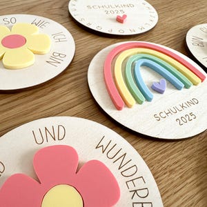 Könnte beinhalten: Vier Holzrundlinge mit verschiedenen Designs. Der erste Rundling hat eine gelbe Blume mit pinker Mitte und den Text "So wie ich bin". Der zweite Rundling hat einen Regenbogen mit einem lila Herz und den Text "Schulkind 2023". Der dritte Rundling hat eine rosa Blume mit gelber Mitte und den Text "Und wunderbar". Der vierte Rundling hat das Alphabet in einem Kreis mit dem Text "Schulkind 2023".