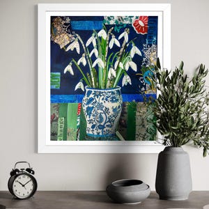 Puede incluir: Una obra de arte enmarcada que presenta un jarrón floral azul y blanco lleno de campanillas de nieve blancas. El fondo es un collage de papel azul, verde y estampado. Un despertador negro, un cuenco gris y un jarrón gris con vegetación están en primer plano.