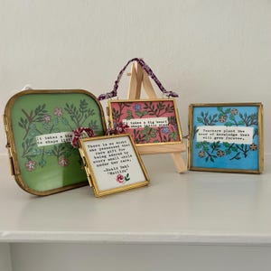 Op de afbeelding: Vier ingelijste kunstwerken met inspirerende citaten. De frames zijn goudkleurig, met bloemmotieven en tekst. Eén frame is groen, een ander roze en een derde blauw. Het vierde frame is wit. De citaten gaan over leraren en kinderen.