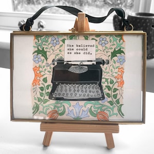 May include: A framed print with a vintage typewriter on a floral background. The print has the quote "She believed she could so she did."