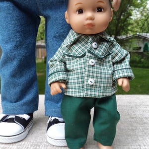 May include: A doll wearing a green and white plaid shirt with matching green pants. The doll is standing in front of a person wearing blue jeans, a red shirt, and black and white sneakers. The doll's clothing is from BibaDollClothes.