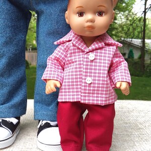 May include: A doll wearing a pink and white checkered shirt with red pants. The doll's outfit includes a collared shirt with white buttons and red pants. The doll is standing in front of a person wearing blue jeans and black and white sneakers.