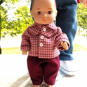 May include: A doll wearing a red and white plaid shirt with white buttons and burgundy pants. The doll is standing in front of a person wearing blue jeans and white sneakers. The website address www.GrannyBibasDollClothes.com is visible at the bottom.