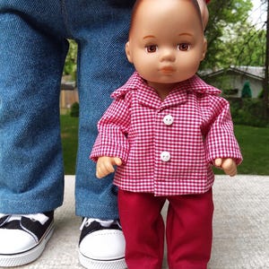 May include: A doll wearing a red and white checkered shirt and red pants. The doll is standing in front of a person wearing blue jeans and black and white sneakers. The doll's outfit has white buttons. The website address www.etsy.com/shop/BibaDollClothes is visible.