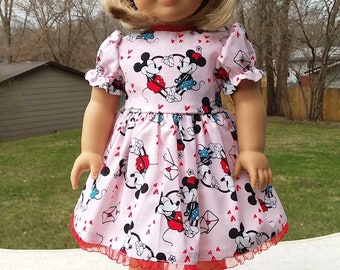 May include: A pink dress with a red and white Mickey Mouse and Minnie Mouse print. The dress has a ruffled hem and is worn by a doll with blonde hair. The doll is wearing red sneakers with white laces.