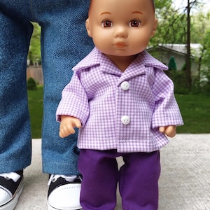 May include: A doll wearing a purple and white checkered shirt with matching purple pants. The doll has brown hair and is standing in front of a person wearing blue jeans and a red shirt. The doll is wearing a button-down shirt.