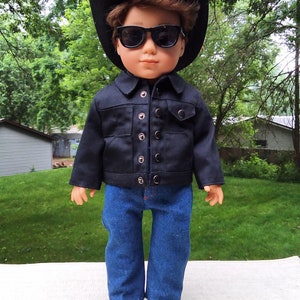 May include: A doll wearing a black denim jacket, blue jeans, a black cowboy hat, and sunglasses. The doll has brown hair and is standing on a white surface.