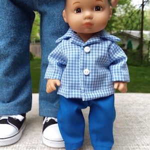 May include: A doll wearing a blue and white checkered shirt with matching blue pants. The doll has brown eyes and is standing in front of a person wearing blue jeans and black and white sneakers.
