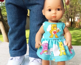 Op de afbeelding: Twee poppen, een draagt een rood jack en een blauwe spijkerbroek, en de andere draagt een blauwe jurk met een Care Bears-print. De pop in de jurk heeft bruin haar en bruine ogen.