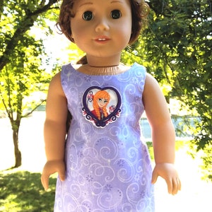 May include: A purple sleeveless dress with a white and purple floral pattern. The dress features a heart-shaped applique with an image of a girl with long red hair. The text "www.GrannyBibasDollClothes.com" is visible at the bottom of the image.