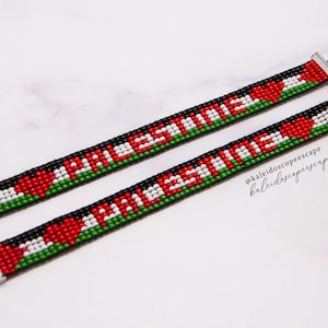 Puede incluir: Dos pulseras de cuentas con la palabra "PALESTINA" en cuentas blancas sobre fondo negro, con detalles de corazón rojo y verde. Las pulseras tienen cierres y extensiones de cadena plateados.