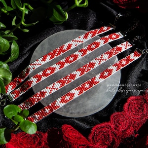 May include: Five beaded chokers with red and white geometric patterns are displayed on a grey stone. The chokers are arranged in a row, with a black background and green foliage in the upper left corner. Red lace is visible in the lower right.