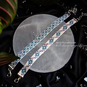 May include: Two beaded bracelets with a geometric pattern in white, blue, and purple. The bracelets have silver-colored clasps and are displayed on a light gray circular surface against a black background.