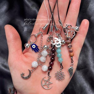 May include: A set of seven charm phone straps with various charms, including a hamsa hand, an evil eye, a crescent moon, a star, an Om symbol, a flower, and a wing. The charms are attached to black cords and feature different colored beads.