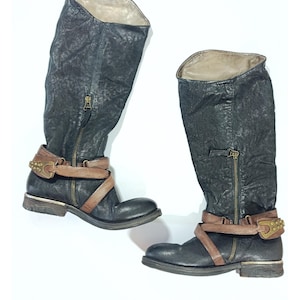 May include: A pair of knee-high boots in distressed black leather with a textured finish. Each boot features a side zipper, a light brown leather strap wrapped around the ankle, and a metallic buckle with small studs on the outer side.