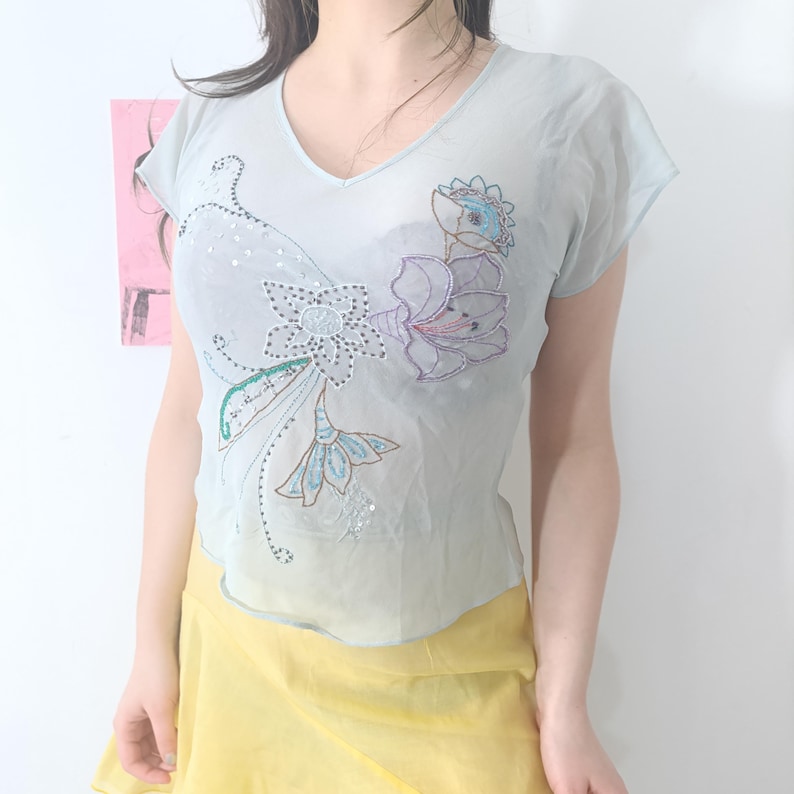 Puede incluir: Top transparente azul claro con cuello en V y mangas cortas. El top presenta bordados florales en tonos azules, morados y verdes, con detalles de lentejuelas. En la parte inferior de la imagen se ve una falda amarilla.