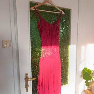 May include: A sleeveless fuchsia dress featuring a V-neckline and thin spaghetti straps. The bodice is adorned with intricate dark beadwork in a swirling pattern, while the skirt consists of layered, vertical fabric strips that create a fringe effect.