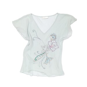 Puede incluir: Una blusa de cuello en V azul claro con mangas cortas. Presenta un dise&ntilde;o floral bordado con detalles blancos, rosas y azules, adornado con peque&ntilde;os detalles brillantes. La tela parece ligera y fluida.