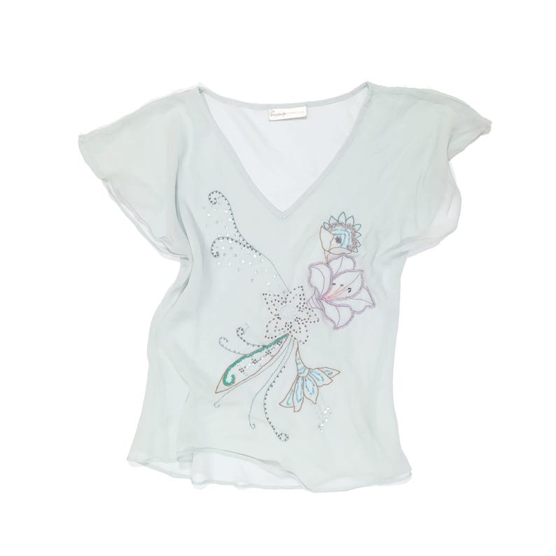 Puede incluir: Una blusa de cuello en V azul claro con mangas cortas. Presenta un dise&ntilde;o floral bordado con detalles blancos, rosas y azules, adornado con peque&ntilde;os detalles brillantes. La tela parece ligera y fluida.
