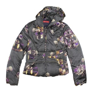 Könnte beinhalten: Eine schwarze Steppjacke mit einem lila und goldenen Druck von Zahnrädern und anderen mechanischen Teilen. Die Jacke hat eine Kapuze und einen Reißverschluss. Das Label "eight sixty" ist auf der Jacke sichtbar.