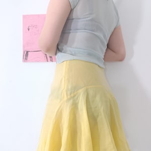 Puede incluir: Una blusa transparente azul claro con mangas cortas y una falda amarilla acampanada. La falda tiene un dobladillo con volantes y ribete amarillo. El conjunto se encuentra sobre un fondo blanco con una pintura abstracta rosa.