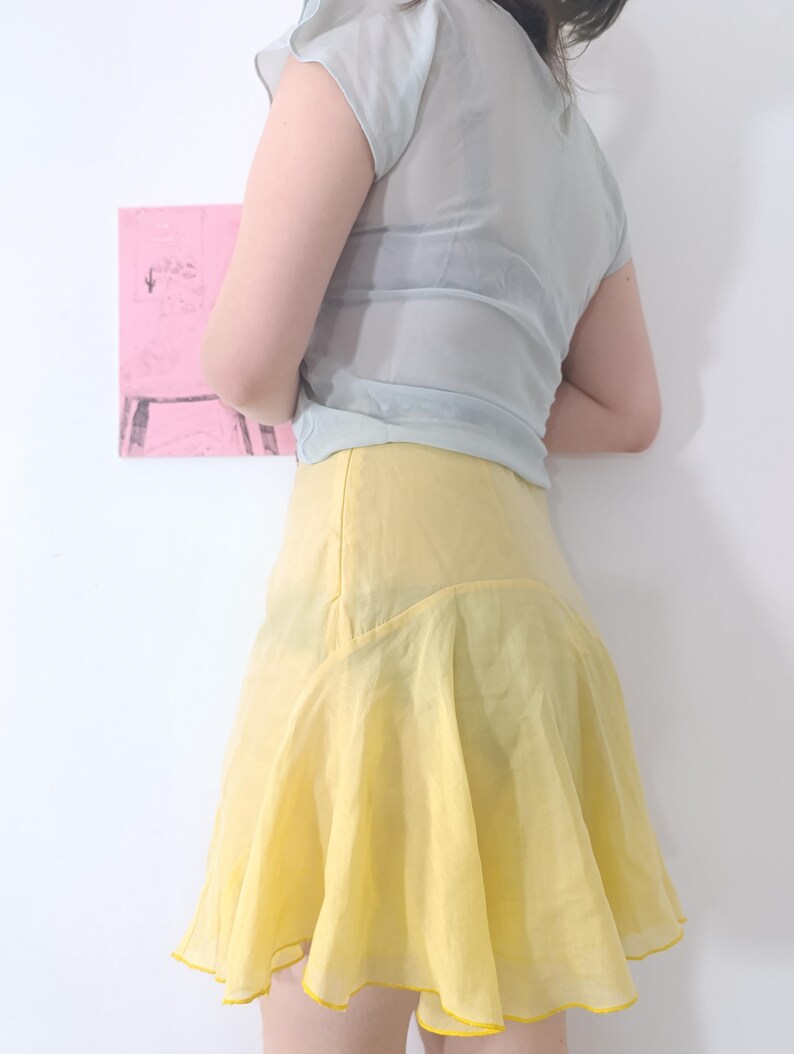 Puede incluir: Una blusa transparente azul claro con mangas cortas y una falda amarilla acampanada. La falda tiene un dobladillo con volantes y ribete amarillo. El conjunto se encuentra sobre un fondo blanco con una pintura abstracta rosa.