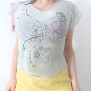 Puede incluir: Una blusa azul claro de manga corta con cuello en V, adornada con dise&ntilde;os florales bordados en tonos azules, morados y plateados. La tela transparente est&aacute; adornada con lentejuelas. La blusa se combina con una falda amarilla.