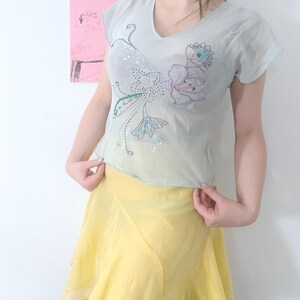 Puede incluir: Una blusa azul claro con cuello en V con bordados florales y lentejuelas. La blusa se combina con una falda amarilla. El conjunto es ligero y parece estar hecho de una tela transparente.
