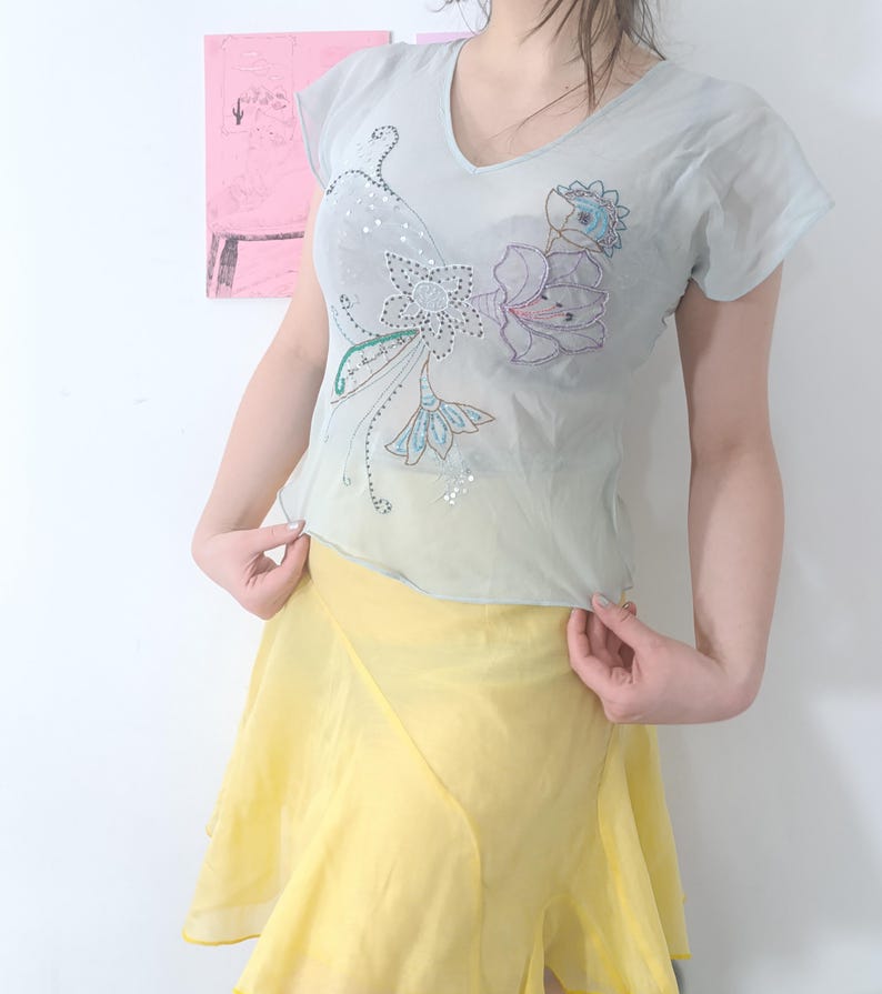 Puede incluir: Una blusa azul claro con cuello en V con bordados florales y lentejuelas. La blusa se combina con una falda amarilla. El conjunto es ligero y parece estar hecho de una tela transparente.