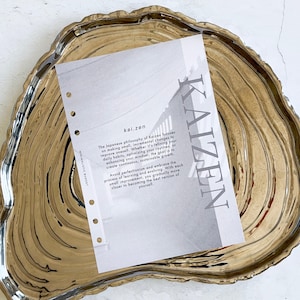 May include: A gold-toned, organic-shaped tray holds a white paper with the word "KAIZEN" in large, vertical text. The paper also includes the text "kai.zen" and a description of the Japanese philosophy of Kaizen.