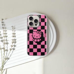 May include: A phone case with a pink and black checkered pattern and a black Hello Kitty graphic. The case is on a white plate with a sprig of dried flowers to the left.