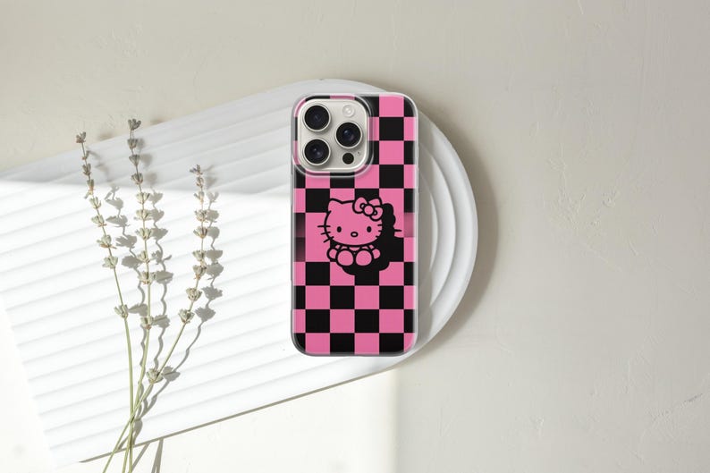 May include: A phone case with a pink and black checkered pattern and a black Hello Kitty graphic. The case is on a white plate with a sprig of dried flowers to the left.
