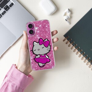 May include: A pink glitter phone case featuring a cartoon cat character with a pink bow and dress. The phone case is held in a person's hand, with a laptop, notebook, and earbuds in the background.