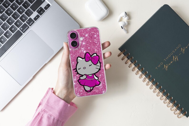 May include: A pink glitter phone case featuring a cartoon cat character with a pink bow and dress. The phone case is held in a person's hand, with a laptop, notebook, and earbuds in the background.