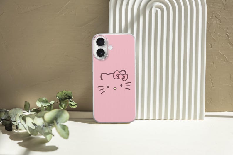 May include: Pink phone case featuring a black outline of a cat face with a bow. The phone case is set against a neutral background with a white arched structure and a sprig of greenery.