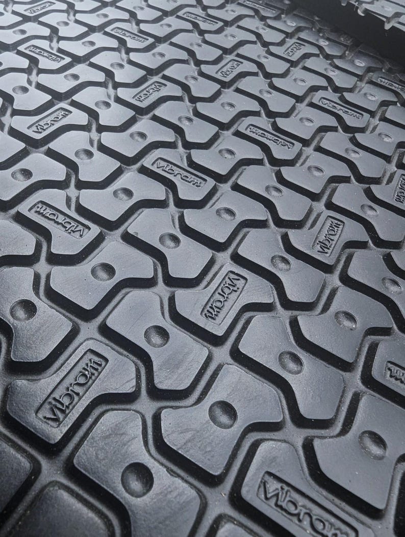 May include: Close-up of a black rubber surface with a repeating pattern of raised, textured squares. The squares have a raised, rounded edge and a small, round indentation in the center. The word "Vibram" is embossed in white on each square.